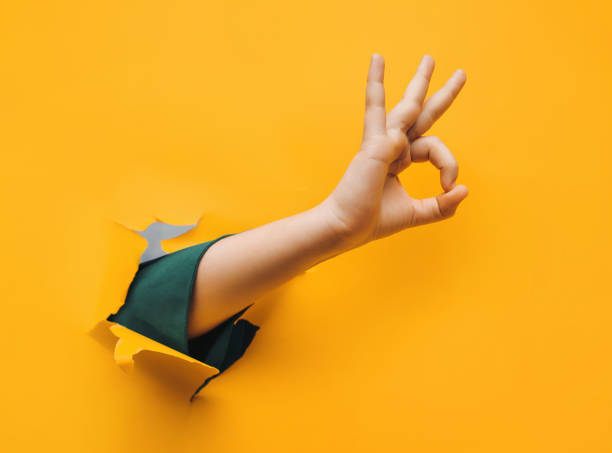 Hand OK sign. The teenage hand came out into the hole of yellow paper and shows symbol of Okay. stock photo