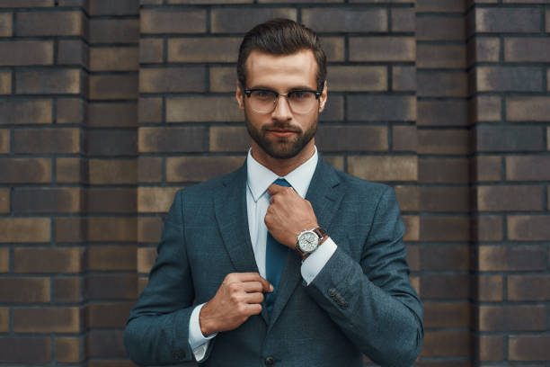 istock Perfect look. Confident businessman in full suit adjusting his tie and looking at camera while standing against brick wall 1170677156