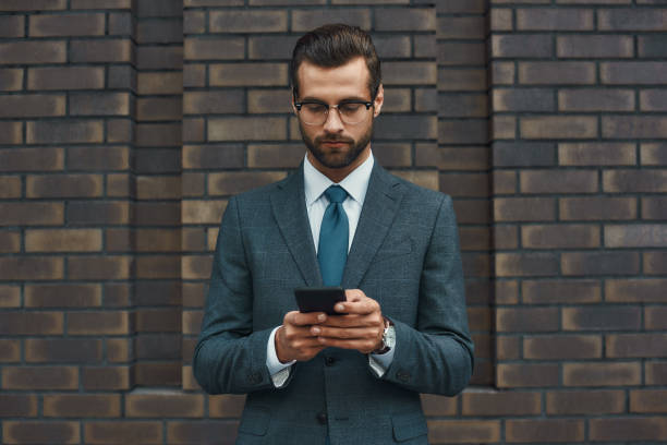 istock Business message. Handsome businessman in formal wear and eyeglasses using smart phone while standing against brick wall 1170677330