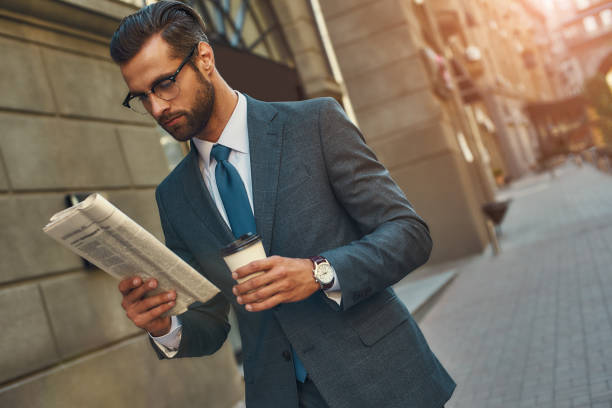 若いハンサムなひげを生やしたビジネスマンは、新聞を読み、屋外を歩きながらコーヒーを飲みながら眼鏡をかけた - young adult reading newspaper the media ストックフォトと画像