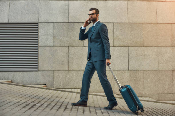 istock Always in touch. Side view of handsome bearded man in suit pulling his luggage and talking by phone with client while walking outdoors 1170677419