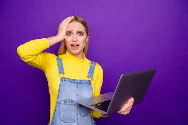 Photo of Portrait of impressed girl hold gadget touch her head wear yellow turtleneck denim jeans overalls isolated over purple violet background