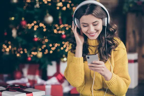 Photo of Photo of charming beautiful wavy curly haired girl listening to music wearing yellow pullover staring at telephone screen