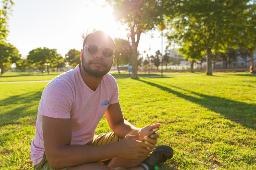 Relaxed Latin guy using phone in park. Focused man in casual and sunglasses sitting on grass, holding smartphone and looking at camera. Wireless connection concept