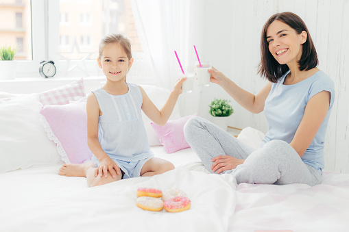 Horizontal shot of pretty female and her daughter clink glasses with cocktail in bed, have breakfast in bedroom, eat delicious doughnuts, look happily at camera. Young mother and her kid at home