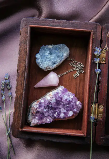 Witch tools inside beautiful old wood box. Rose quartz pendulum, natural amethyst and celestite crystal clusters. Dry lavender flowers on dark purple cloth. Alternative lifestyle concept.