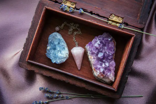 Witch tools inside beautiful old wood box. Rose quartz pendulum, natural amethyst and celestite crystal clusters. Dry lavender flowers on dark purple cloth. Alternative lifestyle concept.