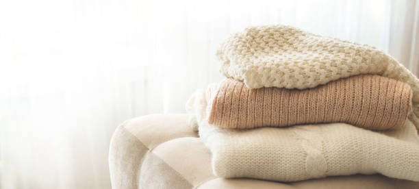 Stos luksusowych swetrów z alpaki w neutralnych odcieniach na miękkim krześle na jasnym tle.