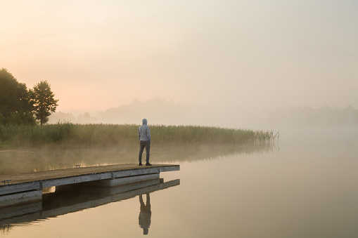 Young man standing alone on wooden footbridge and staring at lake. Mist over water. Foggy air. Early chilly morning. Back view.