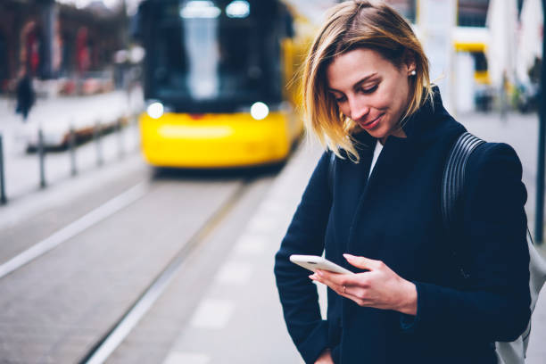 Smiling young woman checking table of city traffic online on web page using smartphone,positive hipster girl satisfied with app for paying for electric transport via cellphone waiting for tram sonriente joven mujer revisando la tabla de tráfico de la ciudad en línea en la página web utilizando el teléfono inteligente, chica hipster positiva satisfecha con la aplicación para pagar el transporte eléctrico a través del teléfono celular espe