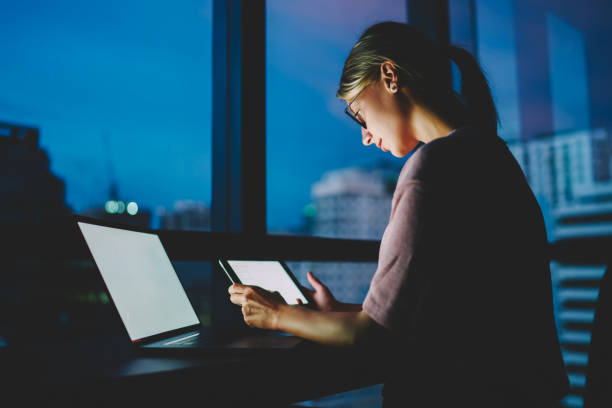 young female freelancer in eyeglasses watching training webinar in webpage on touch pad connected to wireless internet in darkness at home. woman updating software on laptop computer with blank screen - training computer learning computer lab imagens e fotografias de stock