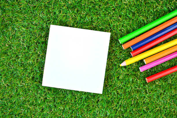 Empty white paper on a green artificial grass and colorful pencils. Minimal composition in flat lay style.
