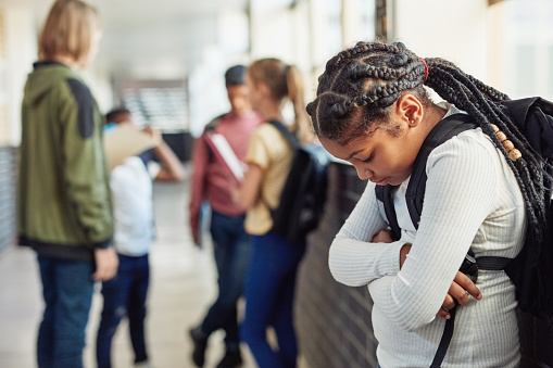 Shot of a young girl looking sad while being excluded from her peers in the hallway of a school