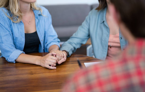 Lesbian couple holding hands during counseling Lesbian couple holding hands during marriage counseling. Young women are visiting mental health professional in office. They are sitting at table. sad gay stock pictures, royalty-free photos & images