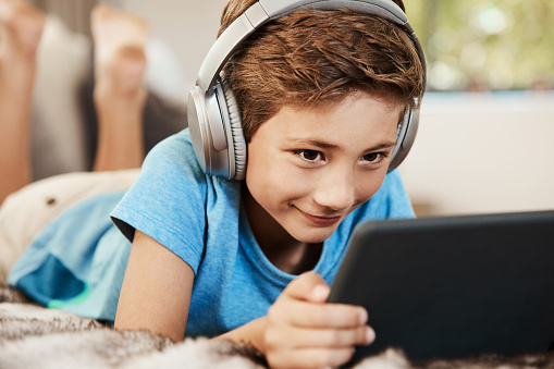 Shot of a cute little boy using a digital tablet with headphones on the sofa at home