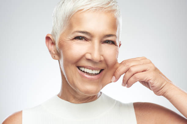 Woman pinching her cheek. Gorgeous smiling Caucasian senior woman with short gray hair pinching her cheek. Beauty photography. face pinch stock pictures, royalty-free photos & images