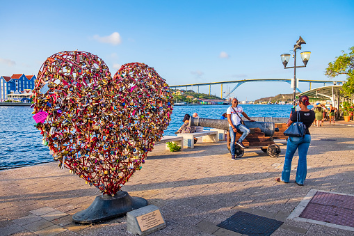 Tourists taking pictures from the beautiful promenade of Punda in Willemstad, the capital city of Curaçao. The heart in foreground is an artistic installation by Carlos Blaaker, dated 2017.