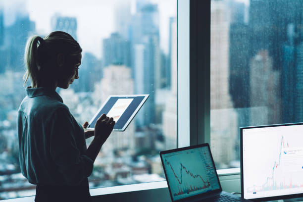 silhouette de femme d'affaires restant dans l'intérieur de bureau près de la fenêtre de gratte-ciel avec le garniture de contact dans des mains. économiste de femme contrôlant la devise de bourse par l'intermédiaire des ressources financières en lig - touchpad photos et images de collection