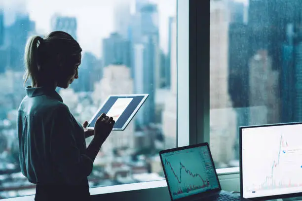 Photo of Silhouette of businesswoman standing in office interior near skyscraper window with touch pad in hands. Woman economist checking stock exchange currency via online financial resources on modern tablet