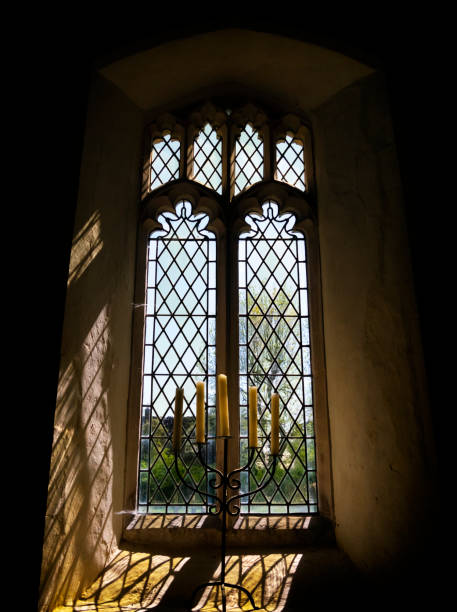 A Perpendicular Style window in a medieval church in Suffolk, Eastern England, on a sunny day. The sun is streaming through the window, reflecting the coloured glass and backlighting the candle holder standing on the sill.