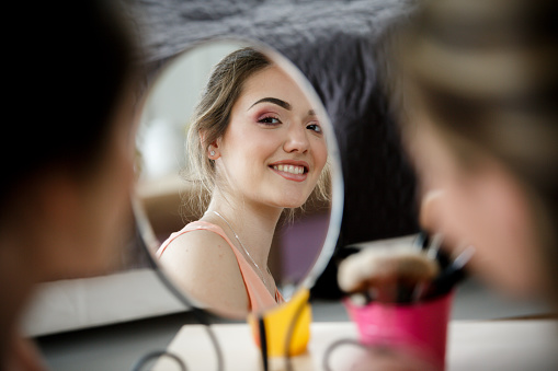 Selective focus of a cheerful young woman looking in the mirror after getting her make up done by a professional make up artist. Seen as reflection in the mirror.