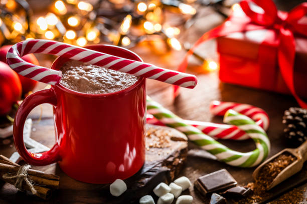 Homemade hot chocolate mug with red and white candy cane on rustic wooden Christmas table Red mug filled with homemade hot chocolate shot on rustic Christmas table. A red and white candy cane is on the hot chocolate mug and two others candy canes are behind the mug. String Christmas lights and a red gift are out of focus on background. Predominant color is red. DSRL studio photo taken with Canon EOS 5D Mk II and Canon EF 100mm f/2.8L Macro IS USM. Candy Cane stock pictures, royalty-free photos & images