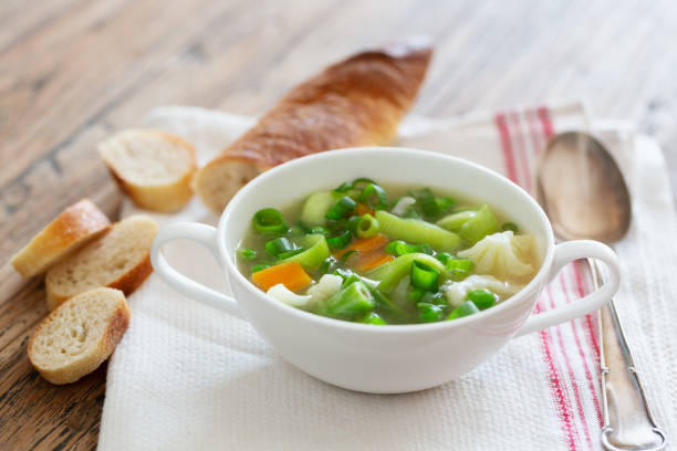 Simple vegetable soup in a white china bowl on rustic wooden background, soup contains beside carrots mostly green vegetables like beans, peas, leek and cauliflower. Besides there are some slices of bread