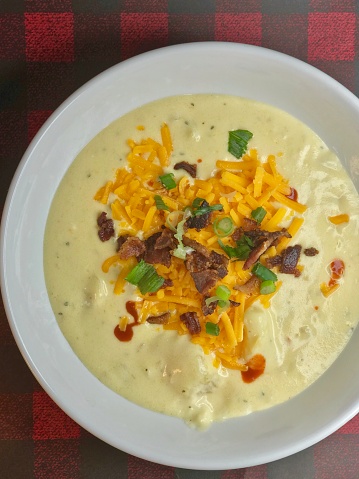 Baked Potato Soup with ultimate toppings forming a warming heart in a  white soup bowl . Background is red flannel place setting for a cozy soup feel. Toppings are cheese, bacon, green onions, and hot sauce scattered on top of the creamy potato soup.