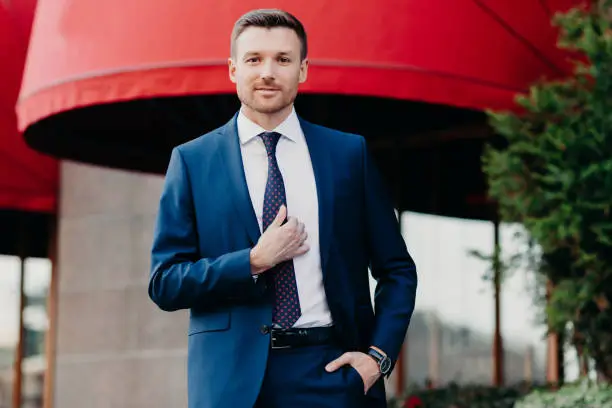 Handsome male company owner dressed formally, stands outside, has confident expression, ready for business meeting. Successful young attractive executive manager poses outdoor near office building