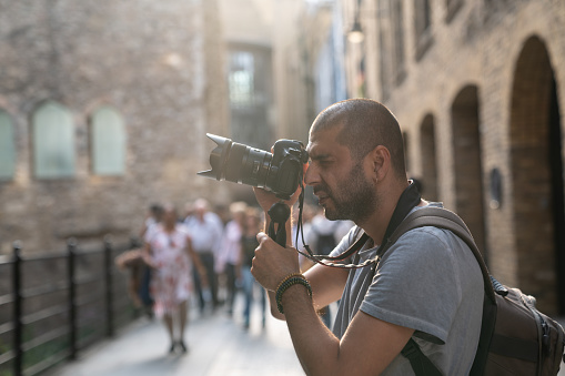 Portraits of adult photographer photographing streets of city of London. Architectural style of city is seen on the background.  He is in casual clothings and has short hair. Selective focus on model while large group of people are seen blurred. Shot under daylight with a full frame mirrorless digital camera.