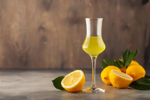Limoncello in glass, sweet Italian lemon liqueur, traditional strong alcoholic drink and lemons on the grey background.