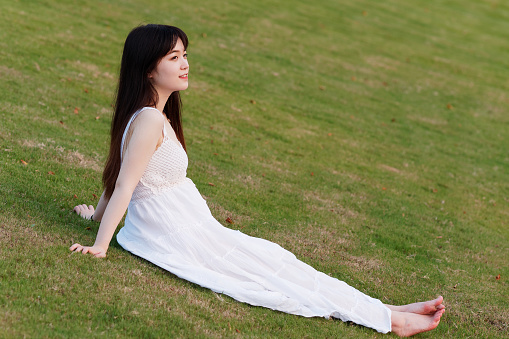 Portrait of beautiful Chinese young woman in white wedding dress sitting on grass field with hand on ground, beauty in summer.