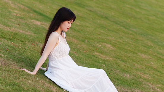 Portrait of beautiful Chinese young woman in white wedding dress sitting on grass field with hand on ground, beauty in summer.