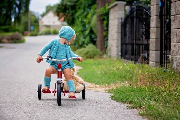 garçon adorable d'enfant en bas âge avec la tenue tricotée, conduisant le tricycle sur une rue tranquille de village - ornamental garden europe flower bed old fashioned photos et images de collection