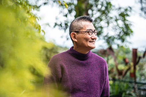 Head and shoulders close-up of smiling senior Chinese man wearing eyeglasses and turtleneck standing in outdoor setting.