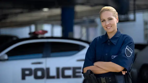 Woman police officer smiling, standing near patrol car with crossed hands, law