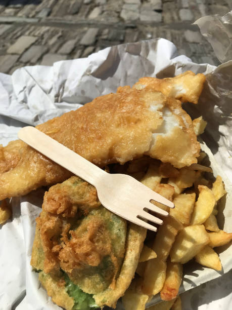Stock photo of takeaway fish and chips in cardboard box with salt and vinegar, unhealthy food takeout, battered cod and deep fried chips as English fish supper ready to eat for lunch at the seaside with fork and mushy pea fritter imagen de pescado para llevar y patatas fritas en caja de cartón con sal y vinagre, comida poco saludable para llevar, bacalao rebozado y patatas fritas fritas como cena de pescado inglés lista para comer para el almuerzo en la costa con tenedor y buñue
