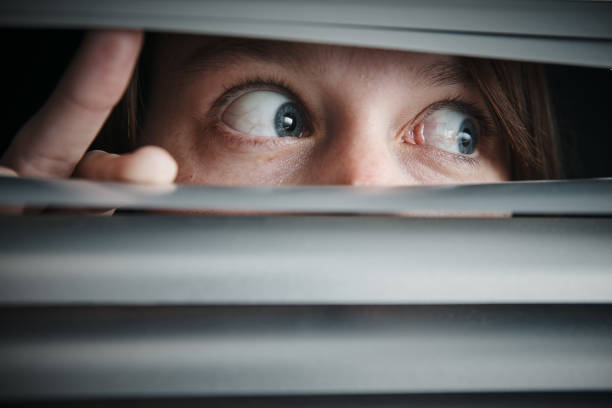 Terrified wide-eyed teenage girl looking through closed blinds A young woman looks to the side through blinds, terrified. phobia stock pictures, royalty-free photos & images
