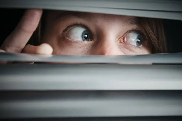 A young woman looks to the side through blinds, terrified.