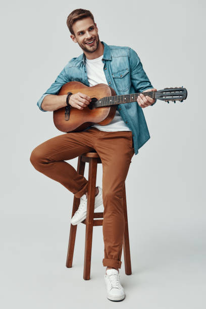 istock Handsome young man smiling and playing guitar while sitting against grey background 1170873526