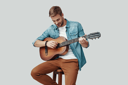 Handsome young man smiling and playing guitar while sitting against grey background