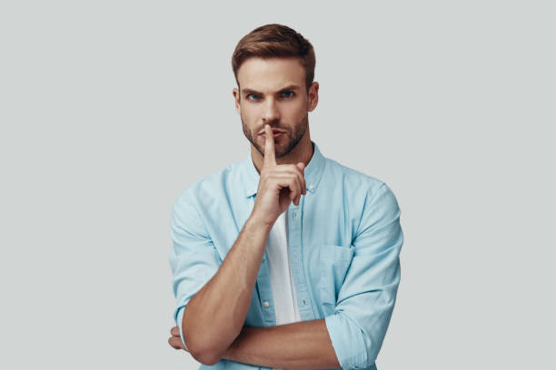 istock Handsome young man looking at camera and keeping finger on lips while standing against grey background 1170875716