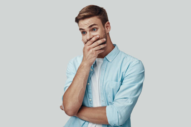 istock Surprised young man covering mouth with hand while standing against grey background 1170875727