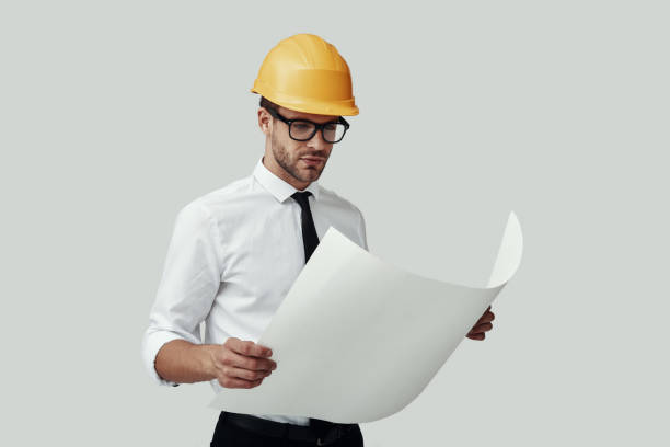 Handsome young man in formalwear looking at blueprint while standing against grey background