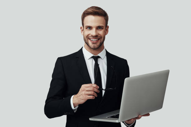 istock Handsome young man in formalwear working using laptop and smiling while standing against grey background 1170877253