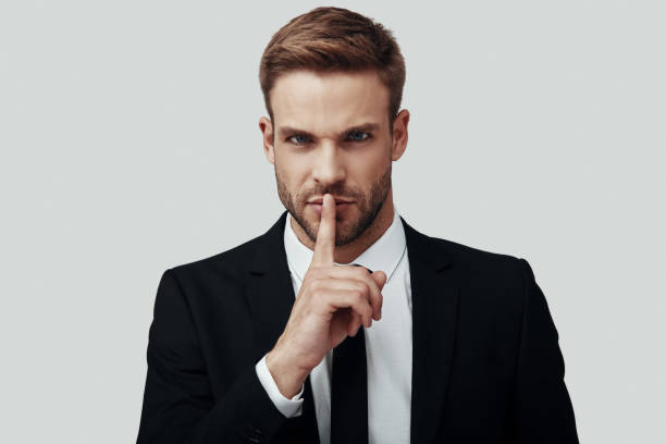 istock Handsome young man in formalwear looking at camera and keeping finger on lips while standing against grey background 1170879426
