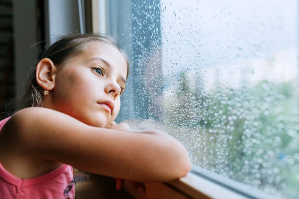 Little sad girl pensive looking through the window glass with a lot of raindrops. Sadness childhood concept image. stock photo