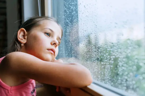 Photo of Little sad girl pensive looking through the window glass with a lot of raindrops. Sadness childhood concept image.