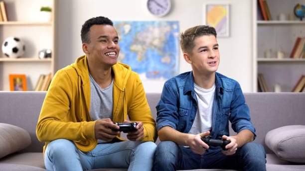 улыбающиеся международные мальчики-подростки с джойстиками, играющими в видеоигры, наркомания - video game friendship teenager togetherness стоковые фото и изображения