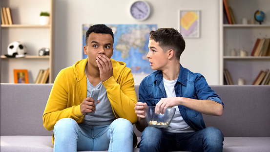 Excited multiracial male teens watching reality show, worrying about heroes